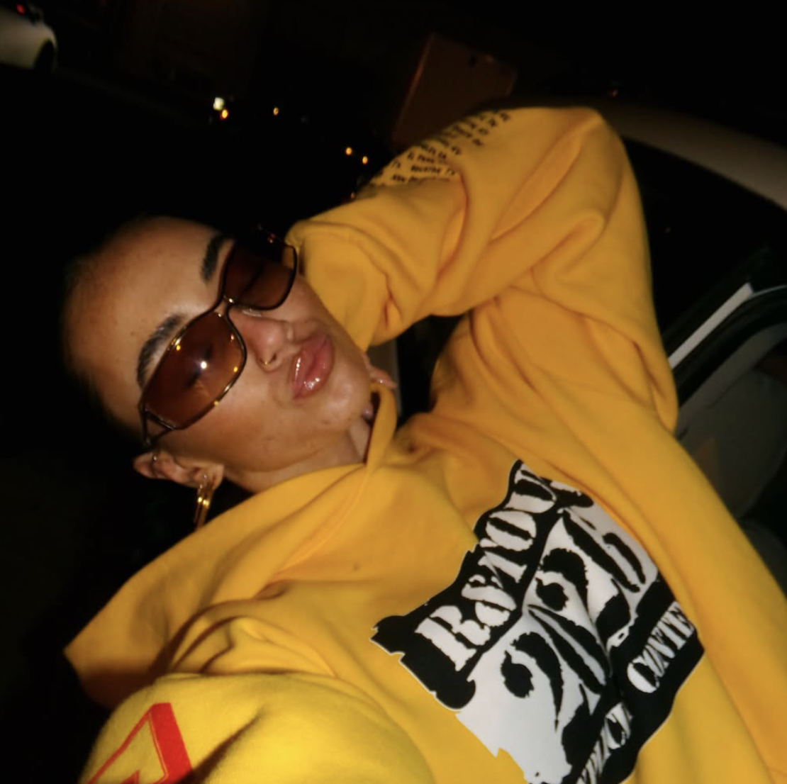 A woman in a yellow hoodie wearing tinted rectangular shades at night